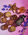 A flat lay of The Star Box items, including assorted spices and ingredients arranged in small bowls on a pink surface. It features an artisan tea strainer, an artistically designed bottle, a glass of the Star Vanilla Ginger Beauty Potion, a star-shaped bowl of dried herbs, and scattered blue flowers.