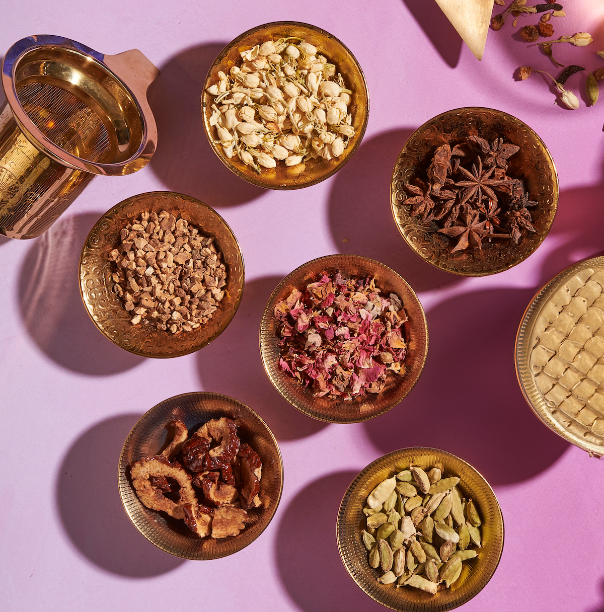 An arrangement of six bowls filled with various dried ingredients on a pink surface. The items include seeds, spices, and dried flowers, perfect for crafting your own loose leaf tea using The Star: Vanilla-Ginger Beauty Potion with Jasmine & Shatavari from Magic Hour. A brass mortar and pestle, along with a triangular cloth bag, are also present in the composition.