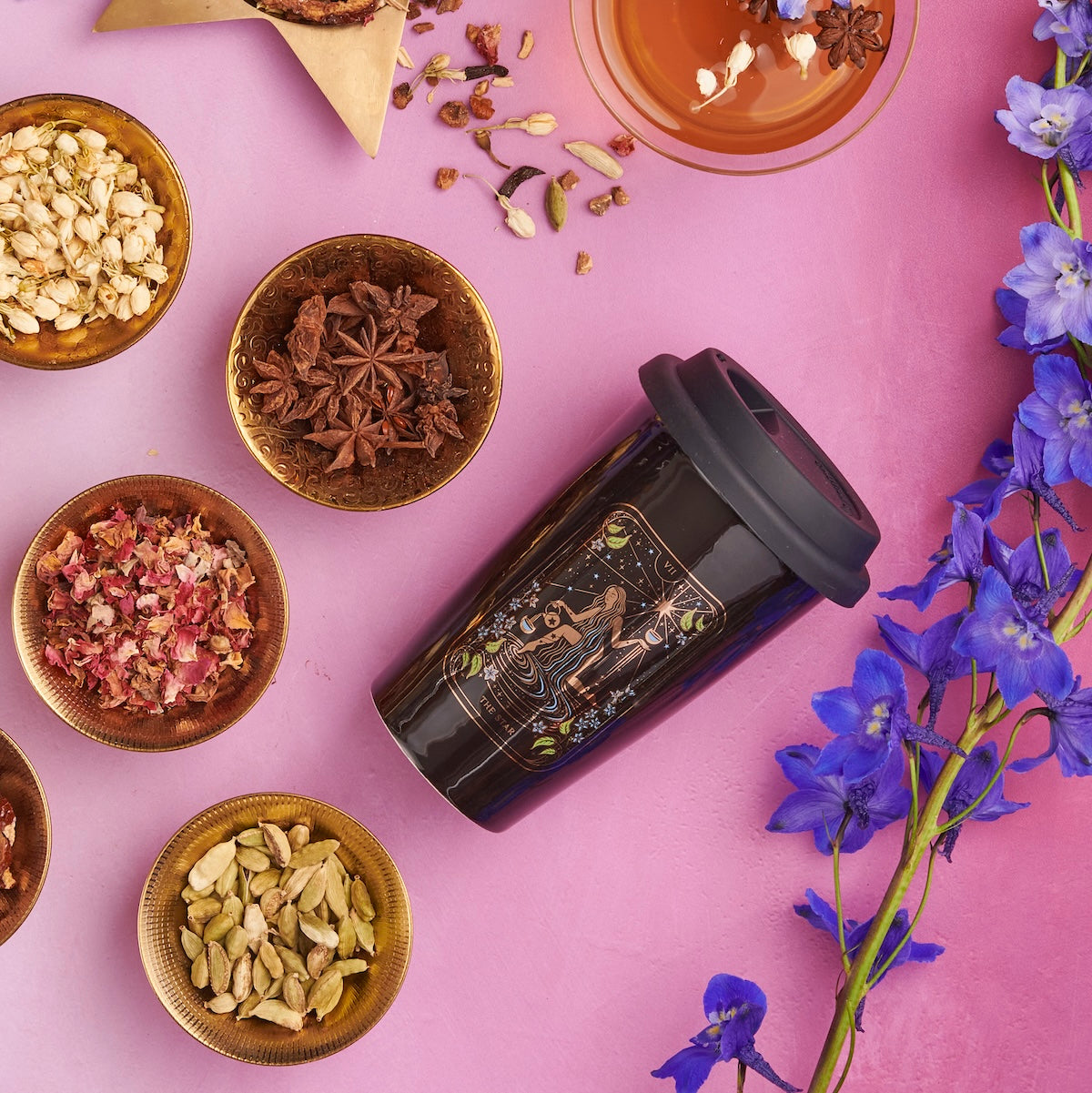 A travel mug with a celestial design is on a pink surface surrounded by bowls containing various dried spices, including star anise, cardamom, and rose petals. Nearby, there's a cup of organic tea and a few pastries, while vibrant blue flowers also adorn the scene.