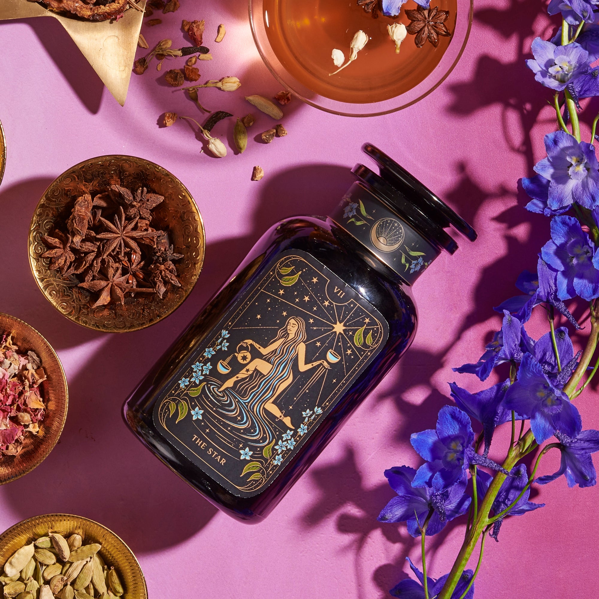 A dark bottle with a label featuring "The Star" tarot card design lays on a pink surface surrounded by various dried herbs in small bowls, a cup of Magic Hour tea, and purple blue flowers. The setup has a mystical and herbalist theme and showcases the Magic Hour product The Star: Vanilla-Ginger Beauty Potion with Jasmine & Shatavari.