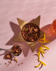 A bowl of Magic Hour’s The Star: Vanilla-Ginger Beauty Potion with Jasmine & Shatavari rests in a star-shaped holder on a pink surface, accompanied by a small glass cup of the tea blend. An ornate spoon holds some loose tea blend with natural flavors. Yellow flower petals are scattered nearby, creating an elegant arrangement.