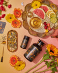 A vibrant flat lay showcases Magic Hour's tea products, "The Sun: Energizing Adaptogenic Herbal Tea with Yaupon, Moringa & Turmeric," in elegant black and gold packaging, surrounded by colorful flowers. A gold tray holds sliced fruits, a tea strainer, and a teacup. Loose Yaupon tea leaves and a golden teaspoon are artfully arranged nearby against a gradient yellow-pink background.