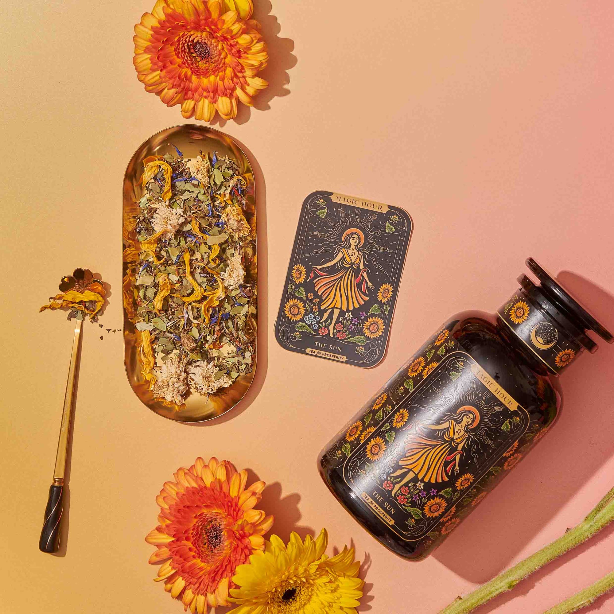 A vibrant flat lay showcases Magic Hour's tea products, "The Sun: Energizing Adaptogenic Herbal Tea with Yaupon, Moringa & Turmeric," in elegant black and gold packaging, surrounded by colorful flowers. A gold tray holds sliced fruits, a tea strainer, and a teacup. Loose Yaupon tea leaves and a golden teaspoon are artfully arranged nearby against a gradient yellow-pink background.