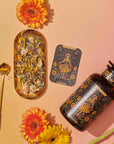 A vibrant flat lay showcases Magic Hour's tea products, "The Sun: Energizing Adaptogenic Herbal Tea with Yaupon, Moringa & Turmeric," in elegant black and gold packaging, surrounded by colorful flowers. A gold tray holds sliced fruits, a tea strainer, and a teacup. Loose Yaupon tea leaves and a golden teaspoon are artfully arranged nearby against a gradient yellow-pink background.