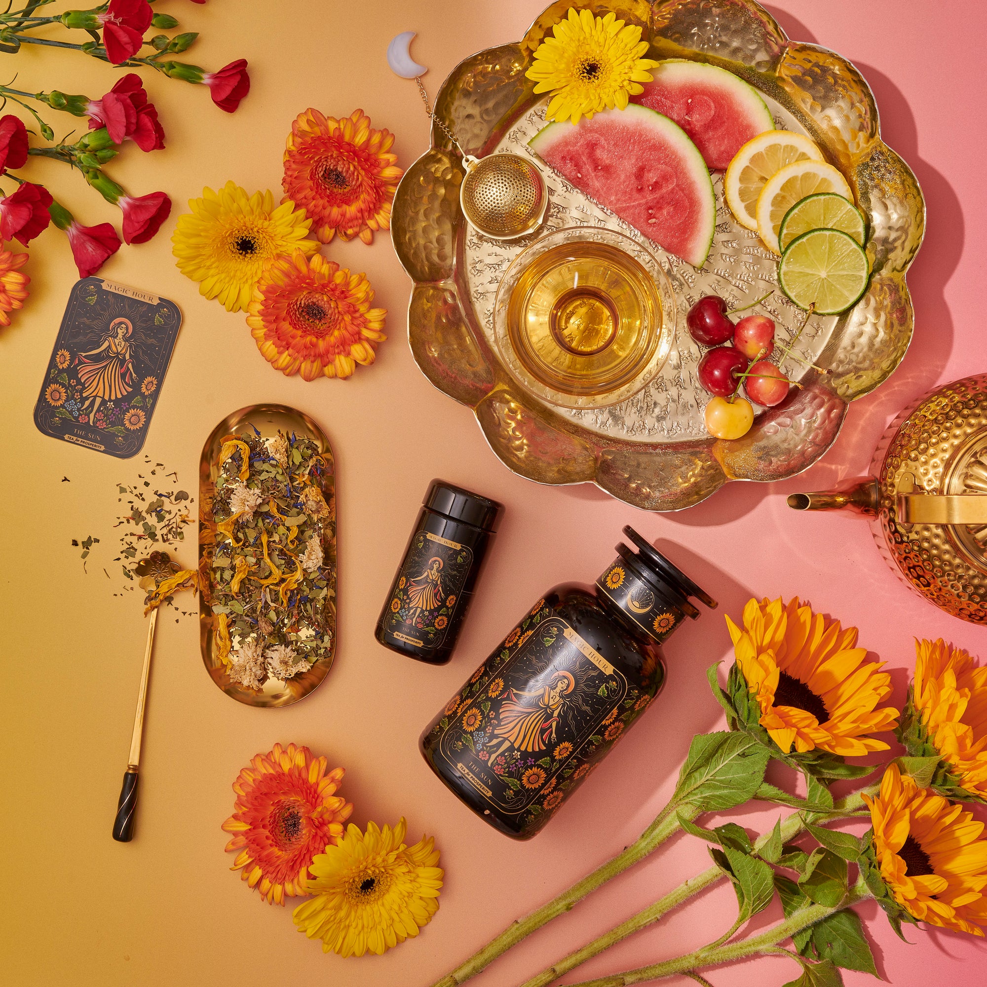 A flat lay featuring The Sun: Energizing Adaptogenic Herbal Tea with Yaupon, Moringa &amp; Turmeric in a floral tin, a golden teapot, two black gold-detailed containers, fresh flowers, and a fruit tray on a pink and yellow background.