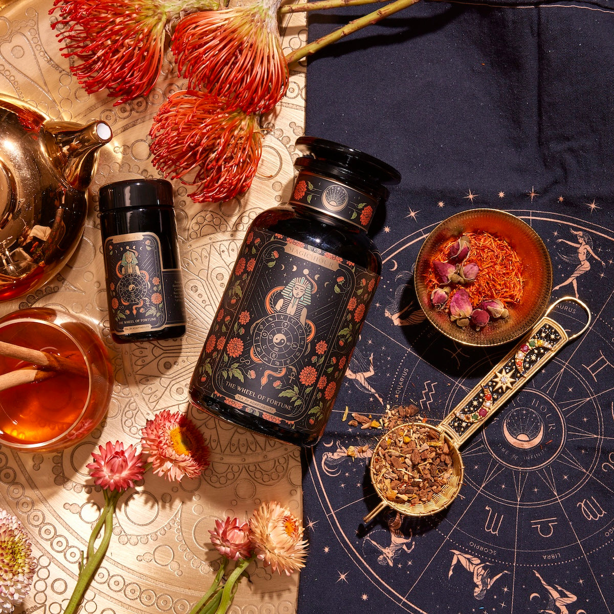 A top-down view showcases The Wheel of Fortune's intricately designed tabletop adorned with astrological and floral motifs. It features a stunning array of herbal products, including both a large jar and a small jar, an elegant tea set, dried flowers, a wooden bowl filled with herbs, a spoon holding loose tea leaves, and dark fabric patterned with zodiac signs.