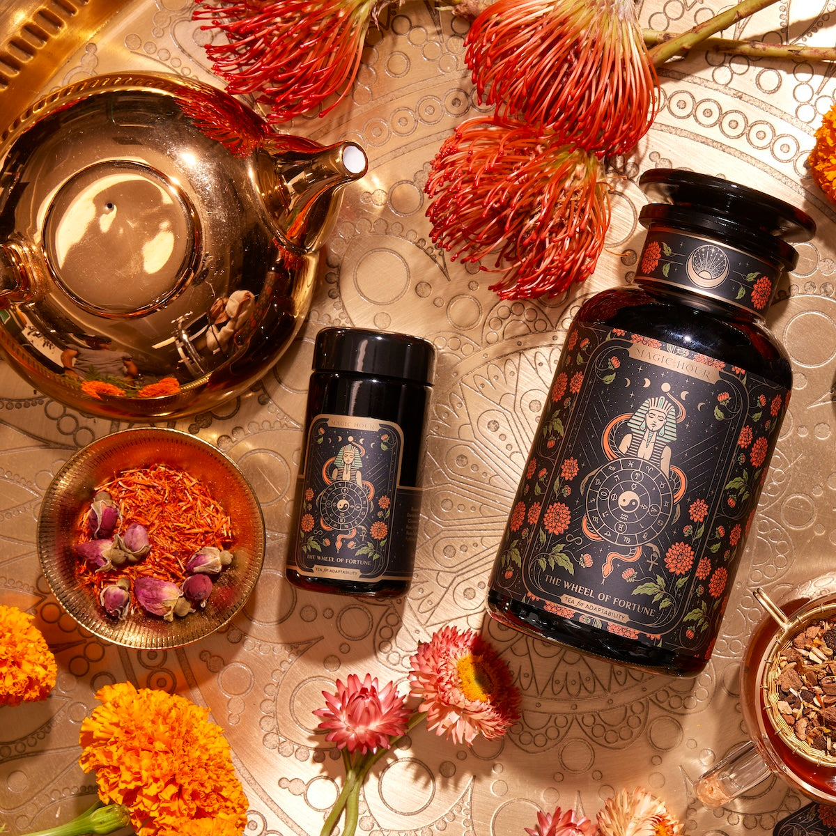 A flat lay photo of "The Wheel of Fortune" tea set on a gold patterned surface. The set includes two dark, decorated tea containers, a gold teapot, and a bowl with red and orange tea leaves. Surrounding the items are bright orange and red flowers.