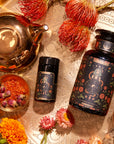 A flat lay photo of "The Wheel of Fortune" tea set on a gold patterned surface. The set includes two dark, decorated tea containers, a gold teapot, and a bowl with red and orange tea leaves. Surrounding the items are bright orange and red flowers.