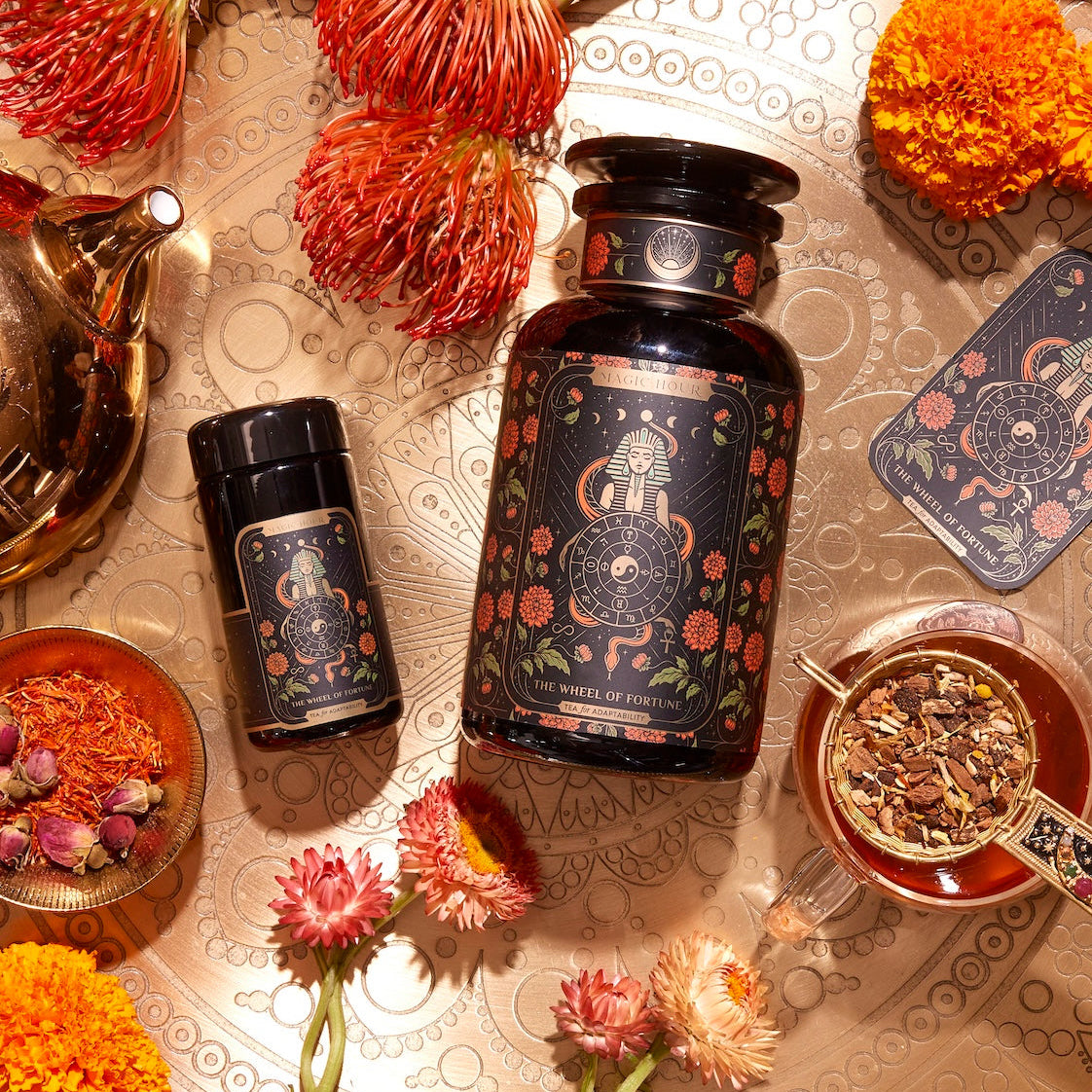 An arrangement featuring a large black jar with a floral design, a smaller matching container, a steaming glass of tea, and a teapot on a decorative surface. Surrounding the items are various vibrant flowers and herbs. Next to the jars lies The Wheel of Fortune tarot card.