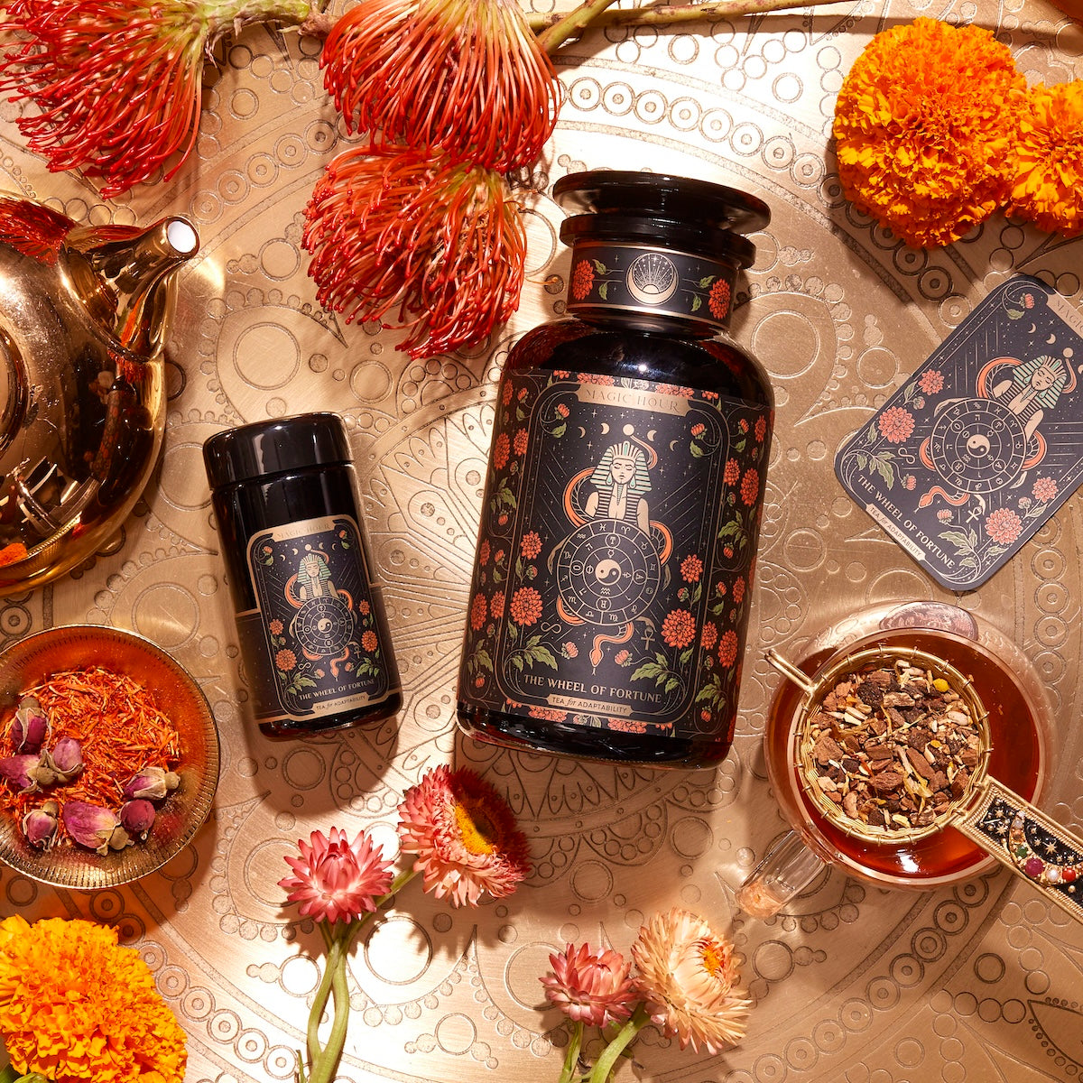 An arrangement featuring a large black jar with a floral design, a smaller matching container, a steaming glass of tea, and a teapot on a decorative surface. Surrounding the items are various vibrant flowers and herbs. Next to the jars lies The Wheel of Fortune tarot card.