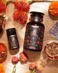 An arrangement featuring a large black jar with a floral design, a smaller matching container, a steaming glass of tea, and a teapot on a decorative surface. Surrounding the items are various vibrant flowers and herbs. Next to the jars lies The Wheel of Fortune tarot card.