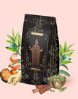 A decorative tea package labeled "The Emperor: Green Chaga Chai with Lion's Mane and Reishi" is surrounded by tea leaves, Gunpowder green tea, cardamom pods, cinnamon sticks, turmeric, and other herbs on a pale pink background—an enticing arrangement that suggests a distinctive blend designed for immune support.
