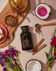 A flat lay image showcasing a bottle of "The Emperor: Green Chaga Chai with Lion's Mane and Reishi," featuring its sleek black design surrounded by fresh flowers, a split coconut, cinnamon sticks, a cup filled with frothy milk, and a wooden tray holding a brass pot along with Gunpowder green tea leaves. A golden teaspoon and scattered tea leaves elegantly complete the scene.