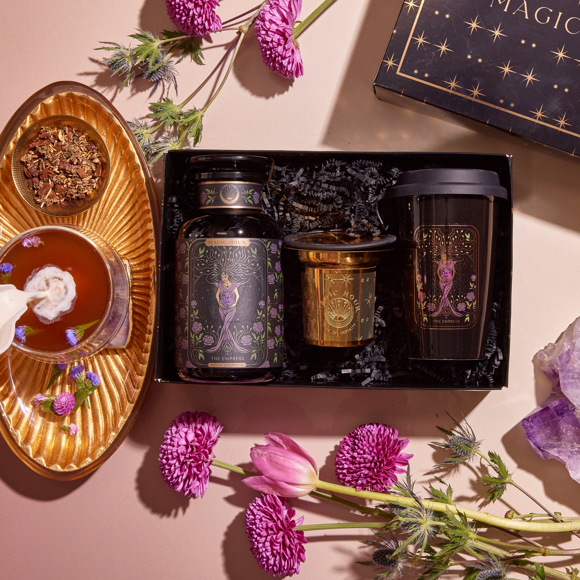 A Tea and Tarot to Go Gift Set features elegantly designed black tea containers and a gold cup, all beautifully packaged in a box adorned with stars. Accompanying the set is a teacup filled with loose leaf tea and a Ceramic Tarot Mug pouring milk. The scene is enhanced by the addition of purple and pink flowers, complemented by a piece of amethyst, creating an ideal presentation for any tea lover or tarot enthusiast.