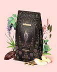 A beautifully decorative tea package featuring mystical, floral artwork rests amid an array of herbs, cocoa, apple slices, and botanicals on a soft pale background—an ideal setting to highlight the soothing qualities of our organic herbal tea. Experience the enchanting blend inside: The Empress—Lavender Currant Shatavari Cocoa Tea of Nurturing Creativity—thoughtfully crafted to inspire serenity and creative balance in your day.