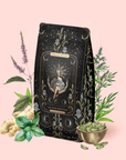 The High Priestess Ginger-Mint-Cardamom Tea Refill Pouch features mystical artwork on a decorative package, surrounded by botanical elements like mint, rosemary, ginger, and cardamom pods against a light pink background. This enchanting presentation perfectly captures the soothing essence of this premium caffeine-free herbal tea.