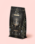 A black and gold refill pouch for The High Priestess Ginger-Mint-Cardamom Tea displays celestial and floral motifs; "Magic Hour" and "The High Priestess" appear above an illustrated mystical figure on the front. The soft pink background evokes the enchanting qualities of this organic, caffeine-free tea.