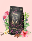A decorative package of The Lovers Hibiscus-Cinnamon-Vanilla Love Potion, adorned with floral and mystical motifs, stands among flowers, cinnamon sticks, vanilla pods, and leaves—creating an enchanting tableau that suggests the balancing herbs inside for women. Set against a soft pink backdrop, this tea blend evokes a sense of calm sensuality.