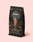 A beautifully designed black refill pouch, adorned with elegant gold, red, and green floral motifs and featuring an illustrated magician holding a wand beneath the "Magic Hour" and "The Magician" labels against a pale pink background, contains The Magician - Elderberry-Ginger-Vanilla Herbal Tea. This organic herbal blend is thoughtfully crafted to provide a variety of wellness benefits in every enchanting sip.