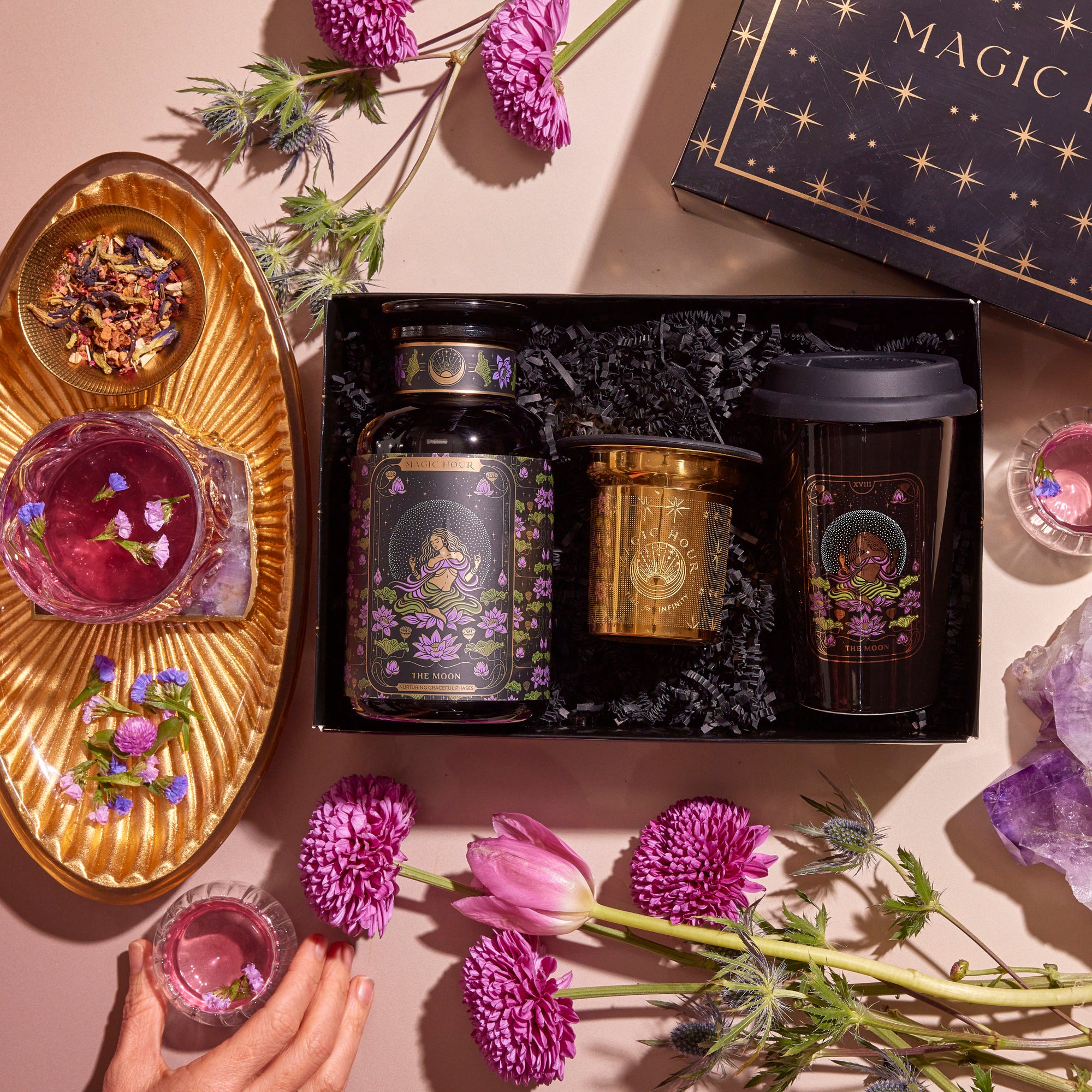 The Tea and Tarot to Go Gift Set embodies mystical elegance, with a selection including tea canisters, a travel mug, and floral design elements. A gold tray holds a cup of purple tea, surrounded by decor inspired by tarot archetypes. Amidst this enchanting setting infused with magic, a hand reaches for the cup.