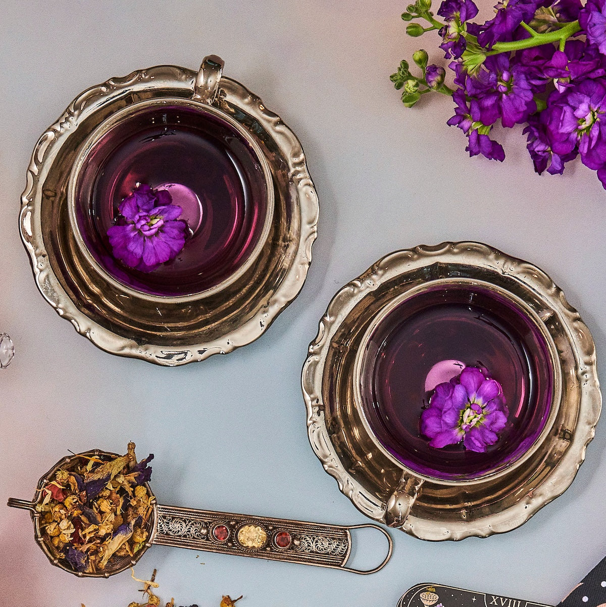 Two ornate silver teacups with thick handles sit on matching saucers, each filled with purple liquid and a floating purple flower. Beside them, a spoon holds dried flowers and herbs. Purple flowers and scattered dried petals decorate the gray surface, creating a Hot Girl Summer Tea Box (Put it on ICE!) experience.