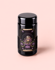 A black cylindrical tea canister features an ornate label depicting a celestial woman seated on lotus flowers beneath a starry sky, adorned with the words “The Moon - Tea for Nurturing Graceful Phases” and “Majestic Cacao Chai.” Infused with butterfly pea flower, this enchanting blend is set against a soft pink background.