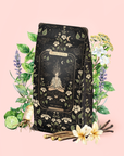 A black and gold patterned tea package labeled "The Oracle: Lemon Lavender Tea for Supporting the Gut-Brain Axis" features a meditating figure surrounded by illustrated herbs, flowers, roots, and spices on a soft pink background. This adaptogenic blend offers a caffeine-free herbal tea experience designed to promote balance and support the gut-brain axis as part of a calming, stress-reducing tea ritual.