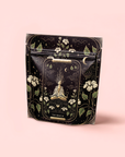 A black and gold package features a meditating woman amid flowers, clouds, stars, and moons. "The Oracle: Lemon Lavender Tea for Supporting the Gut-Brain Axis" hints at an adaptogenic, caffeine-free herbal blend on a soft pink background.