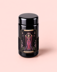A black jar with a decorative label featuring an illustrated woman in a flowing dress, titled “The Queen of Cups: Hibiscus-Vanilla-Berry Tea for Balance & Heart Health,” sits against a light pink background. This special tea blend is thoughtfully crafted to inspire emotional balance and makes a beautiful addition to any herbal tea collection.