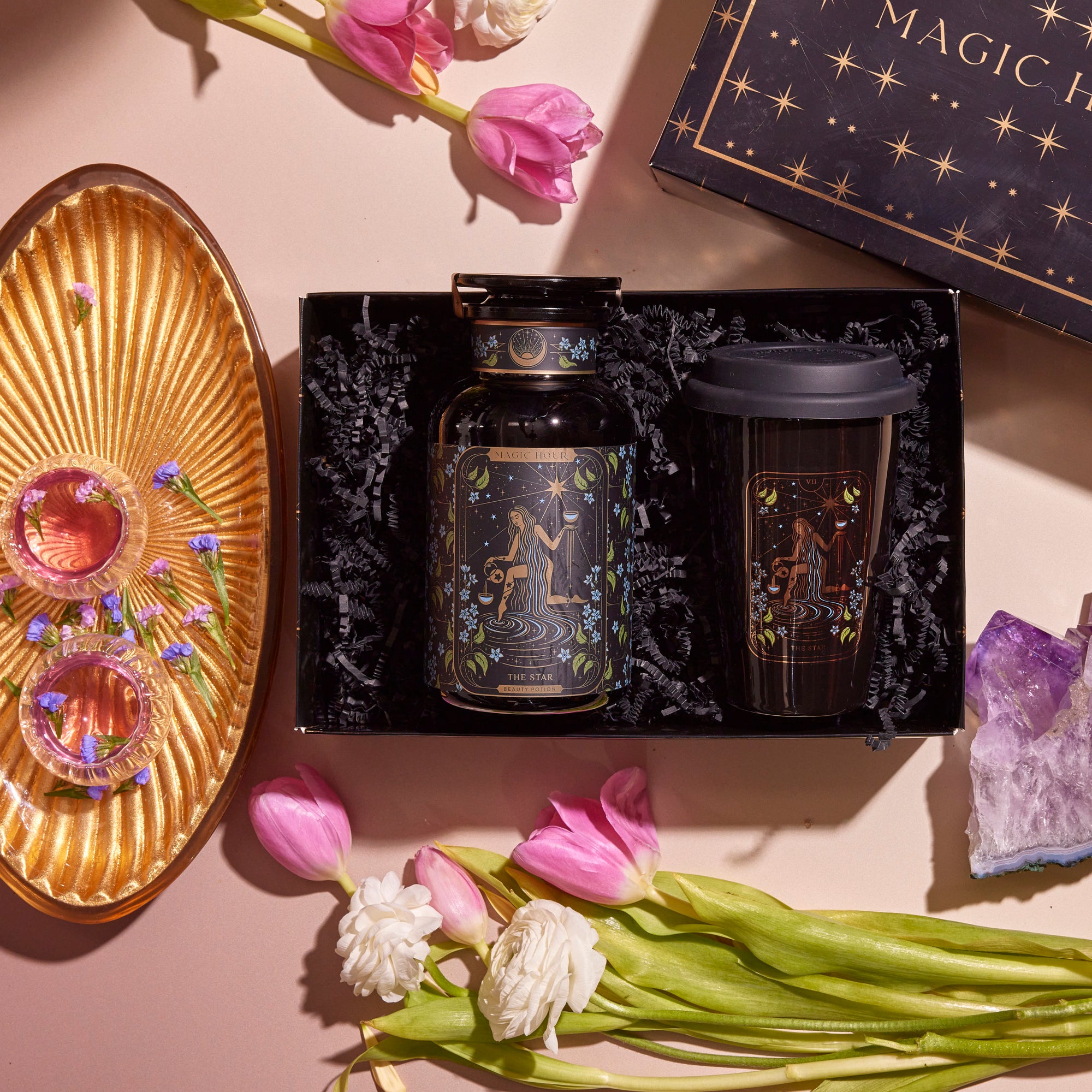 Introducing the captivating Tea and Tarot to Go Gift Set, which includes a beautiful starry jar and a Ceramic Tarot Mug arranged in an elegant black box. It is charmingly complemented by pink tulips, white flowers, an amethyst crystal, a golden tray adorned with gems, and a whimsical book about magic—ideal for those who resonate with the tarot archetype.