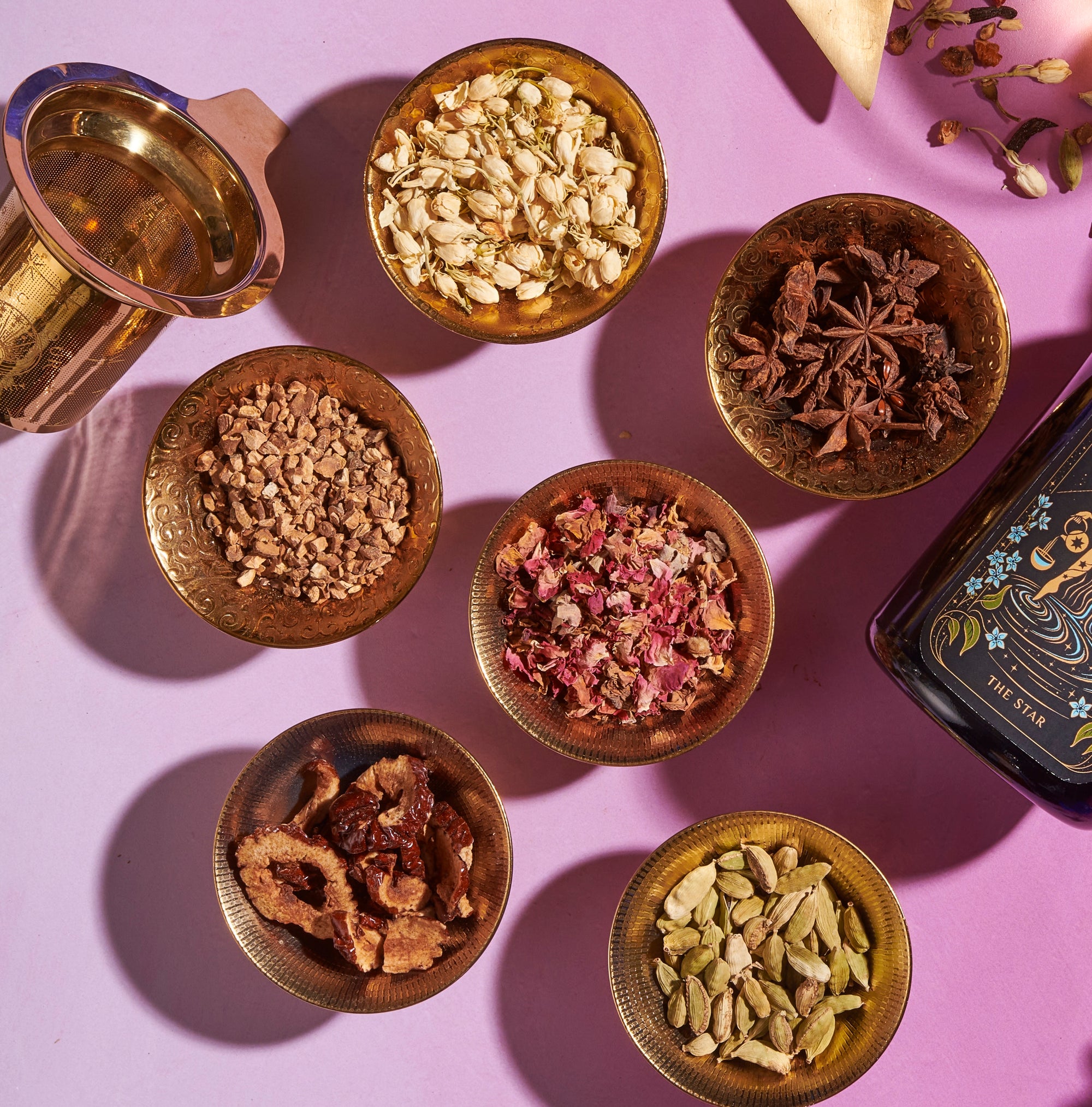 A vibrant display of various dried spices and herbs in small brass bowls, meticulously arranged on a pink surface. The assortment includes cardamom pods, dried flowers, star anise, nutmeg, and other spices. Nearby are a brass mortar and pestle alongside the intricately designed product called The Star Box.