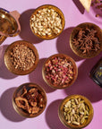 A vibrant display of various dried spices and herbs in small brass bowls, meticulously arranged on a pink surface. The assortment includes cardamom pods, dried flowers, star anise, nutmeg, and other spices. Nearby are a brass mortar and pestle alongside the intricately designed product called The Star Box.