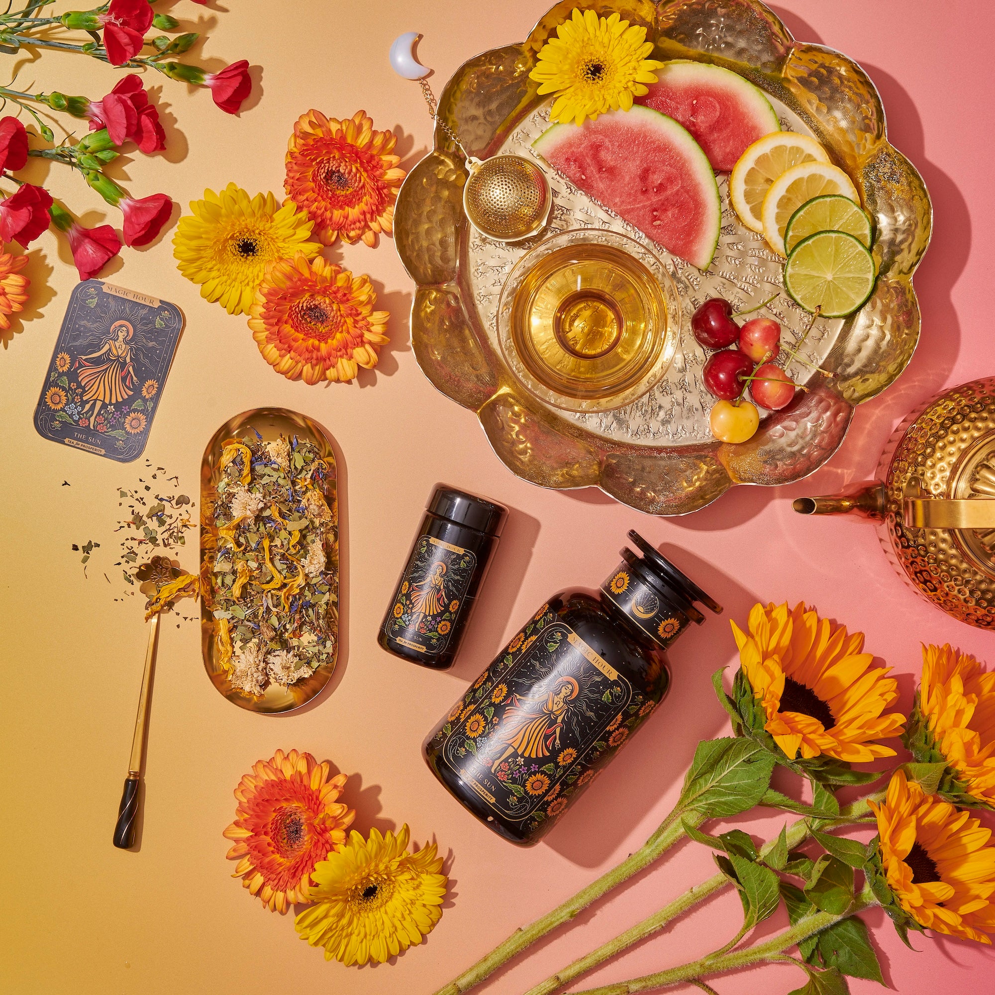 A flat lay image showcasing a vibrant tea setup with floral and fruit accents. Includes a gold teapot, a scalloped tray with watermelon, lime, lemon slices, and cherries, a tea strainer, two black tea containers, a tea blend, a spoon, orange and yellow flowers.