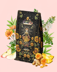 A vibrant package of "The Sun: Energizing Adaptogenic Herbal Tea with Yaupon, Moringa & Turmeric," featuring a sun design, is surrounded by flowers, apple slices, turmeric blend, pineapple, ginger, and green leaves—all arranged on a soft pink background.