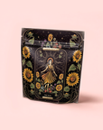 A black package with sunflowers and a central figure in a yellow dress, surrounded by rays and flowers. The label says "THE SUN." Shows off The Sun: Energizing Adaptogenic Herbal Tea with Yaupon, Moringa & Turmeric on a soft pink background.