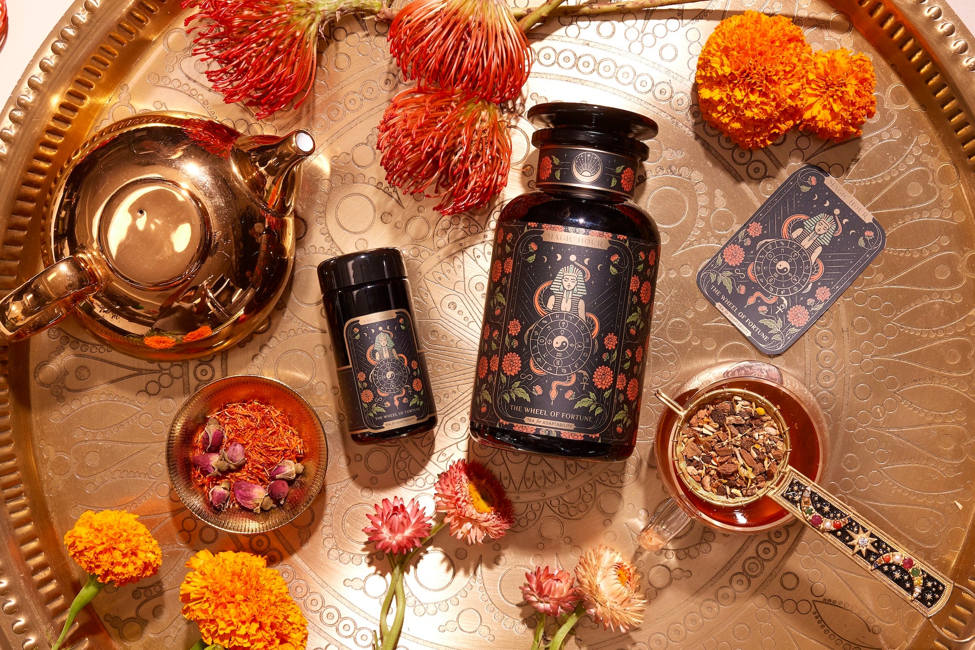 An array of black tea containers with intricate floral designs is set on a golden tray, accompanied by a tea pot, marigold flowers, and a small bowl of loose tea leaves. A spoon with tea sits nearby along with a patterned tea tin and a cup of brewed tea.