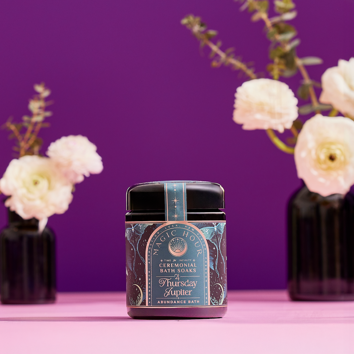 A Ceremonial Bath Salt Soaks - Violet Glass Jar sits on a pink surface with a purple background, flanked by two black vases with white flowers and greenery—ideal for enhancing your self-care ritual.