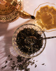 A gold teapot is positioned beside a full, ornate teacup filled with Ti Quan Yin Oolong: Tea of Patient Compassion. In the center, a glass bowl contains loose Ti Quan Yin Oolong: Tea of Patient Compassion leaves with a spoon resting inside. Some loose leaves are scattered around the bowl on the white surface, hinting at their wellness benefits.