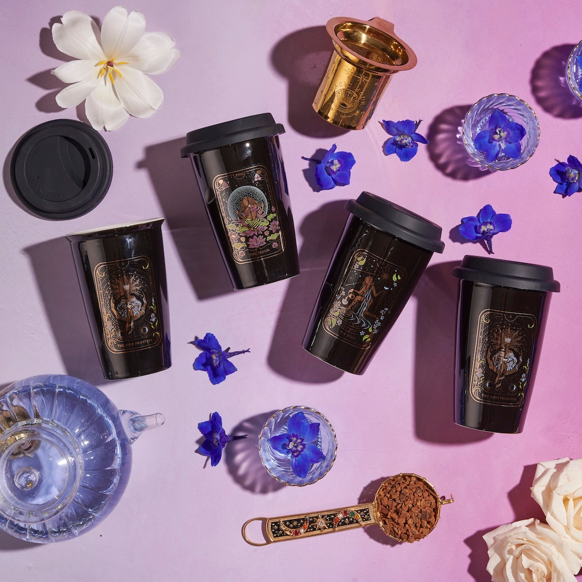 Five black travel mugs with artistic designs are placed on a pink surface, surrounded by blue flowers, a white flower, a clear glass cup, a blue teapot, and a brass tea infuser. A spoon filled with Magic Hour’s The Queen of Cups - Tarot To Go! completes the enchanting scene.