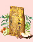 A golden coffee bag adorned with a geometric pattern is surrounded by caramel, pear, apple slices, cinnamon sticks, tea leaves, and green foliage on a light pink background—an inviting scene that beautifully highlights the uplifting and metabolism-boosting benefits of Topaz - Caramel Apple Oolong Tea for Optimism & a Lightness of Being.