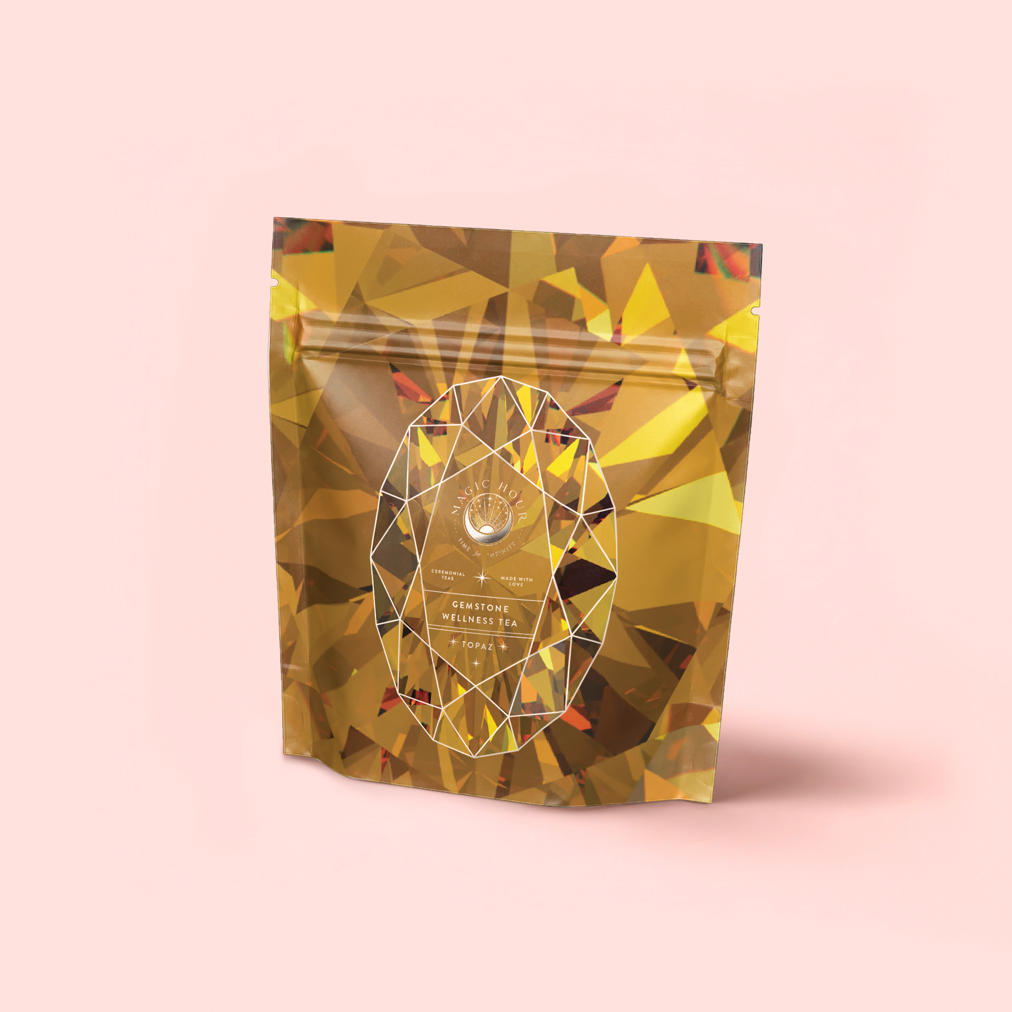 A stand-up pouch with a shiny gold, faceted gemstone design on light pink. The front features a white geometric oval and branding, highlighting Topaz - Caramel Apple Oolong Tea for Optimism & a Lightness of Being.