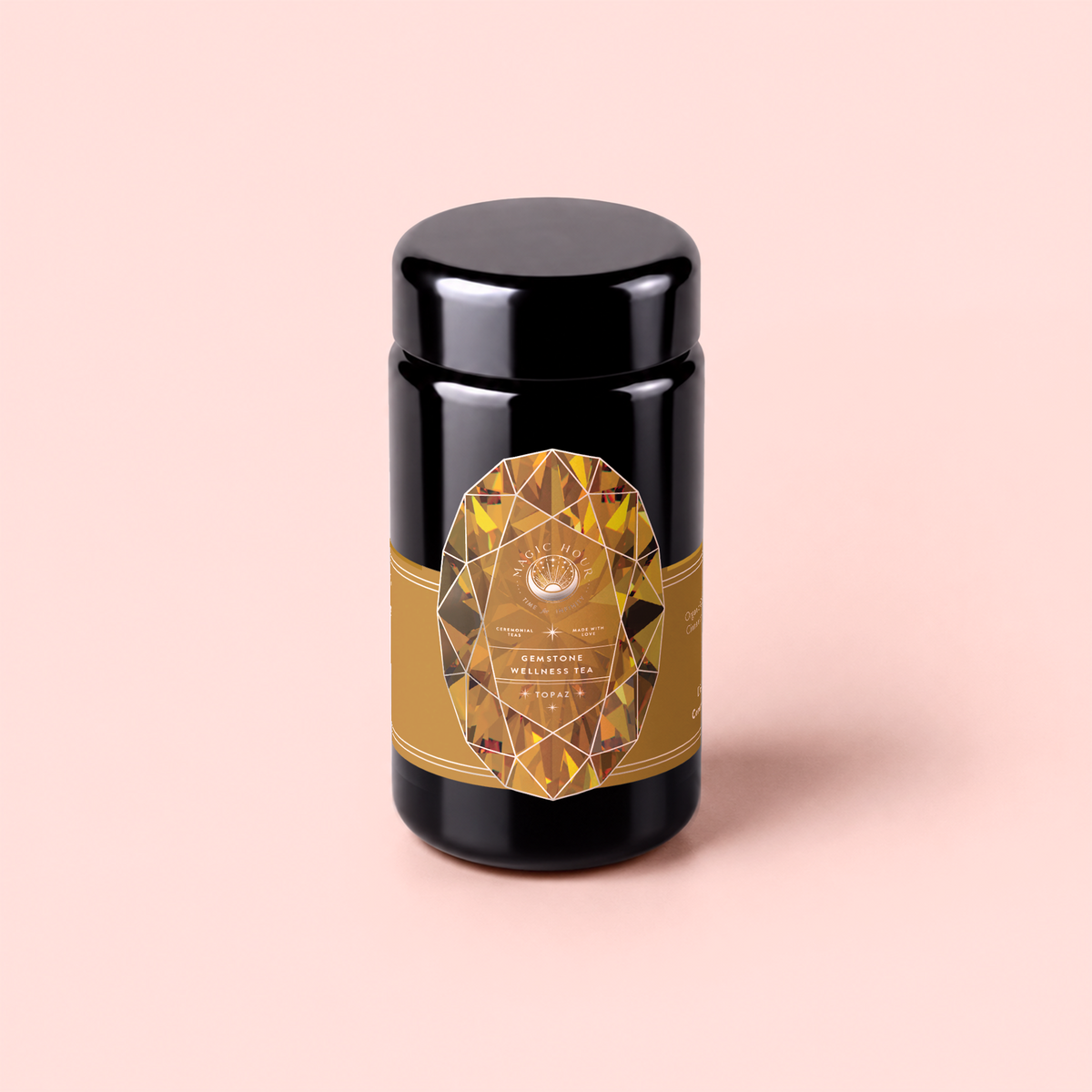 A black cylindrical jar with a gold, gem-patterned label sits on a pale pink background. The label reads “Topaz - Caramel Apple Oolong Tea for Optimism &amp; a Lightness of Being,” hinting at an infusion of organic oolong tea that is renowned for its metabolism-boosting properties and uplifting flavor.