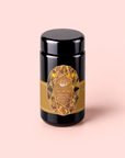 A black cylindrical jar with a gold, gem-patterned label sits on a pale pink background. The label reads “Topaz - Caramel Apple Oolong Tea for Optimism & a Lightness of Being,” hinting at an infusion of organic oolong tea that is renowned for its metabolism-boosting properties and uplifting flavor.