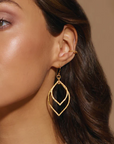 Close-up of a person wearing Magic Hour's Transformed by Light Lotus Linear Earrings with a modern, layered leaf-like design, reminiscent of lotus petals. Their hair is dark brown and wavy, complemented by a small 18kt gold plated ear cuff. The background is a neutral beige color.