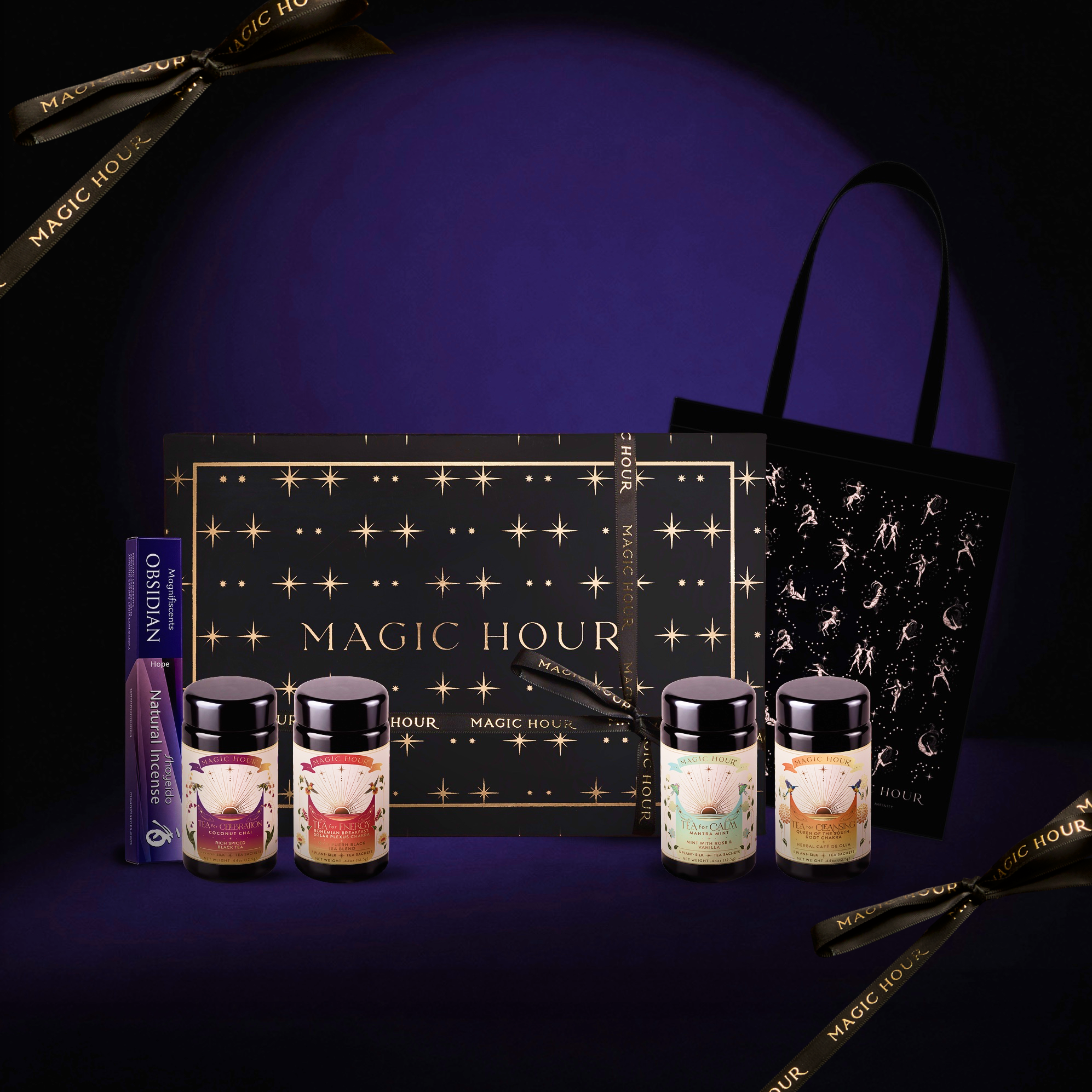 The Traveler’s Spell Kit Box from Magic Hour, featuring tea jars, a box, an astrology tote bag, and a tea tube, is displayed on a dark surface adorned with gold star designs and ribbons.