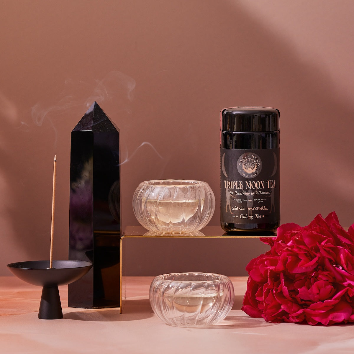 A still life image features a black obelisk, a burning incense stick in a holder, a jar labeled &quot;Triple Moon Tea by Alanis Morissette to Celebrate her Triple Moon Tour!&quot; from Magic Hour, two glass cups releasing steam with magnolia oolong beside them, and a bunch of red roses. All are arranged aesthetically on a neutral background, with jasmine flowers scattered for an added calming effect.