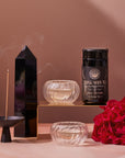 A still life image features a black obelisk, a burning incense stick in a holder, a jar labeled "Triple Moon Tea by Alanis Morissette to Celebrate her Triple Moon Tour!" from Magic Hour, two glass cups releasing steam with magnolia oolong beside them, and a bunch of red roses. All are arranged aesthetically on a neutral background, with jasmine flowers scattered for an added calming effect.