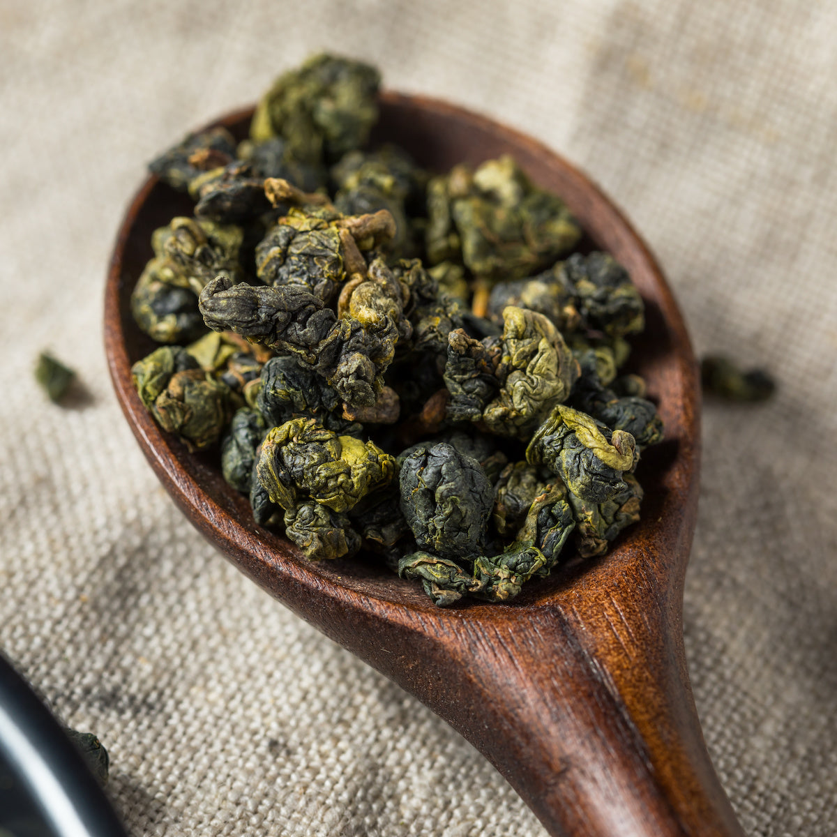 A wooden spoon holds a heap of semi-oxidized oolong tea leaves, displaying a mix of curled and somewhat wavy textures. The spoon rests on a fabric surface, highlighting the rich, dark green and brown tones characteristic of Magic Hour&#39;s Tung Ting Green Dragon Oolong from Taiwan.