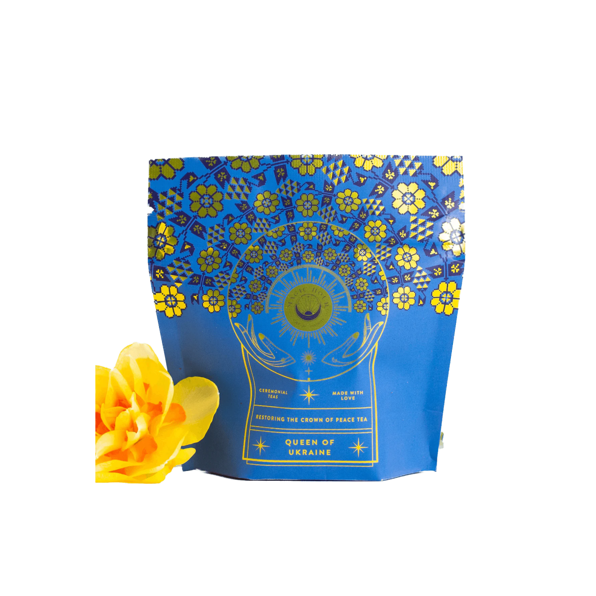 A blue and gold ornate tea packaging labeled "Rest, Relaxation & Sleep Sampler Set" is displayed, featuring a mystical design with floral and celestial motifs. To the left of the package, a bright yellow flower adds a vibrant touch to the composition, enhancing the allure of this Magic Hour Tea.
