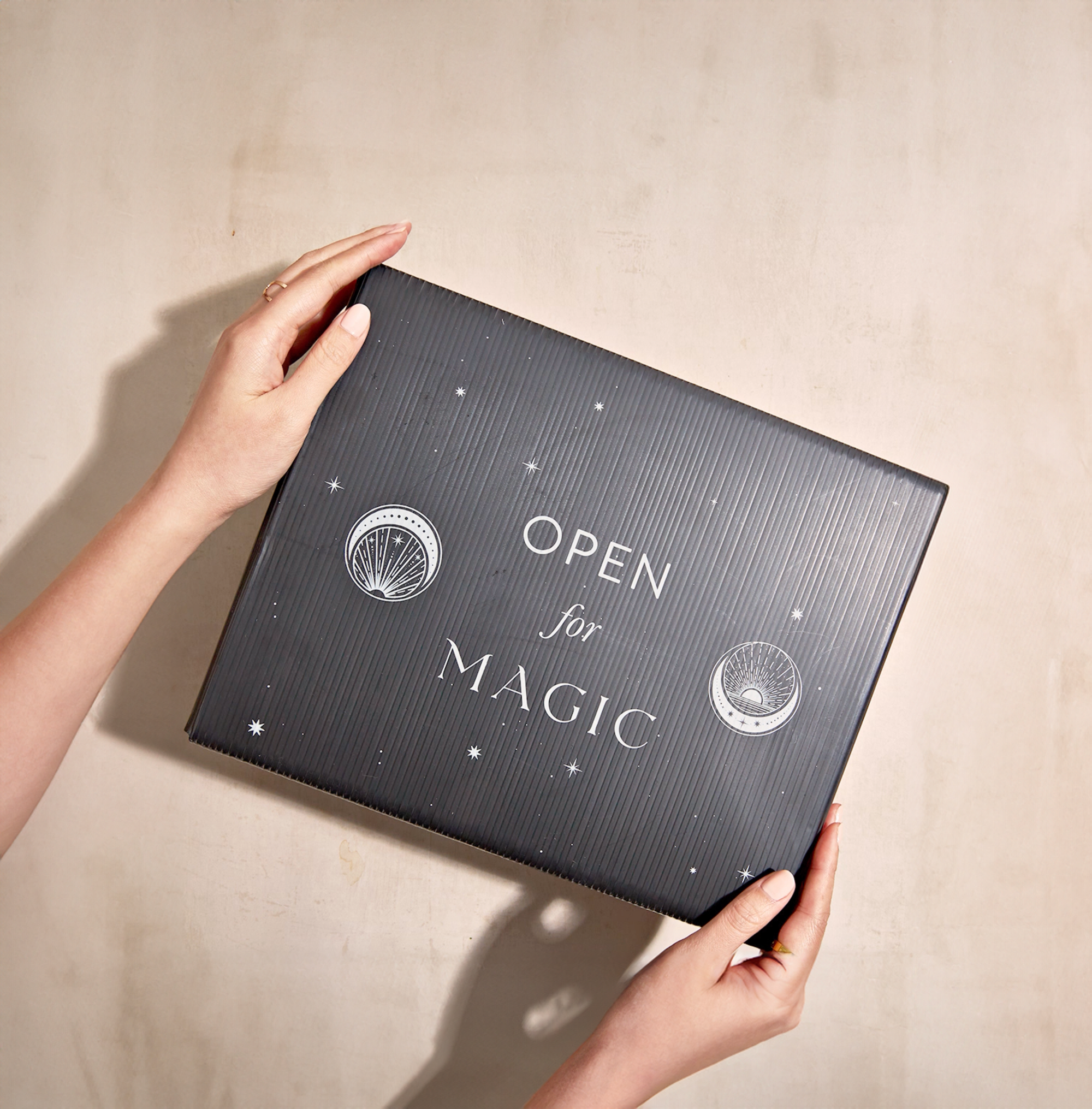 Two hands hold a black gift box emblazoned with white text that reads "Open for Magic" against a beige background. Decorated with tiny stars and circular designs, the box exudes enchantment, suggesting organic botanicals are inside to elevate your wellness routine with the Monthly Magic® First Sips Tea Subscription - Just the Tea!